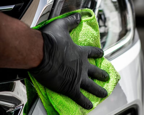 Person cleaning a car window with a green microfiber cloth and black gloves.